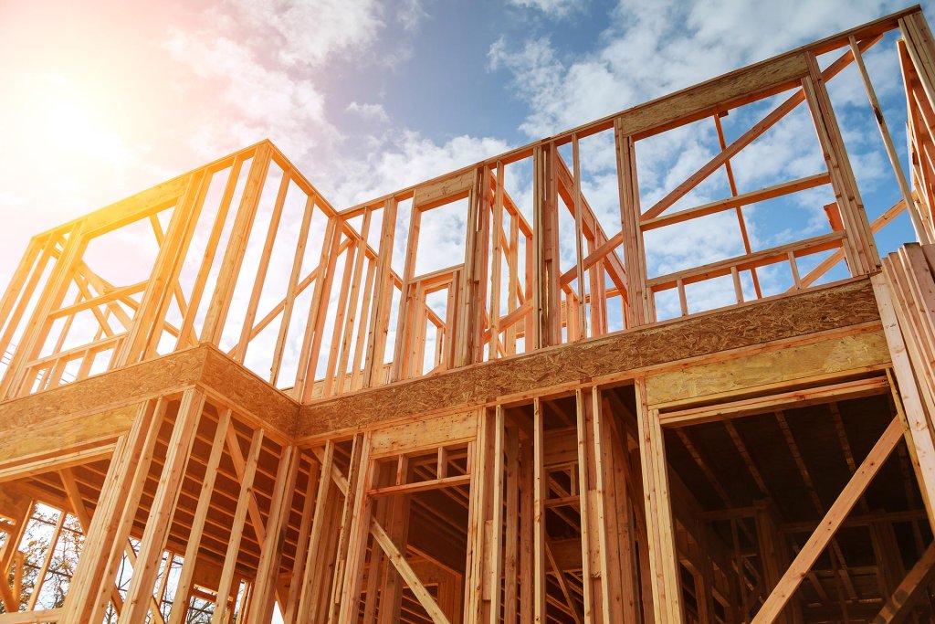 Low-angle view of the structural wood framing of a new two-story home under construction against a bright blue sky with a sun flare. The image showcases professional new construction services, highlighting the initial stages of building a custom home.