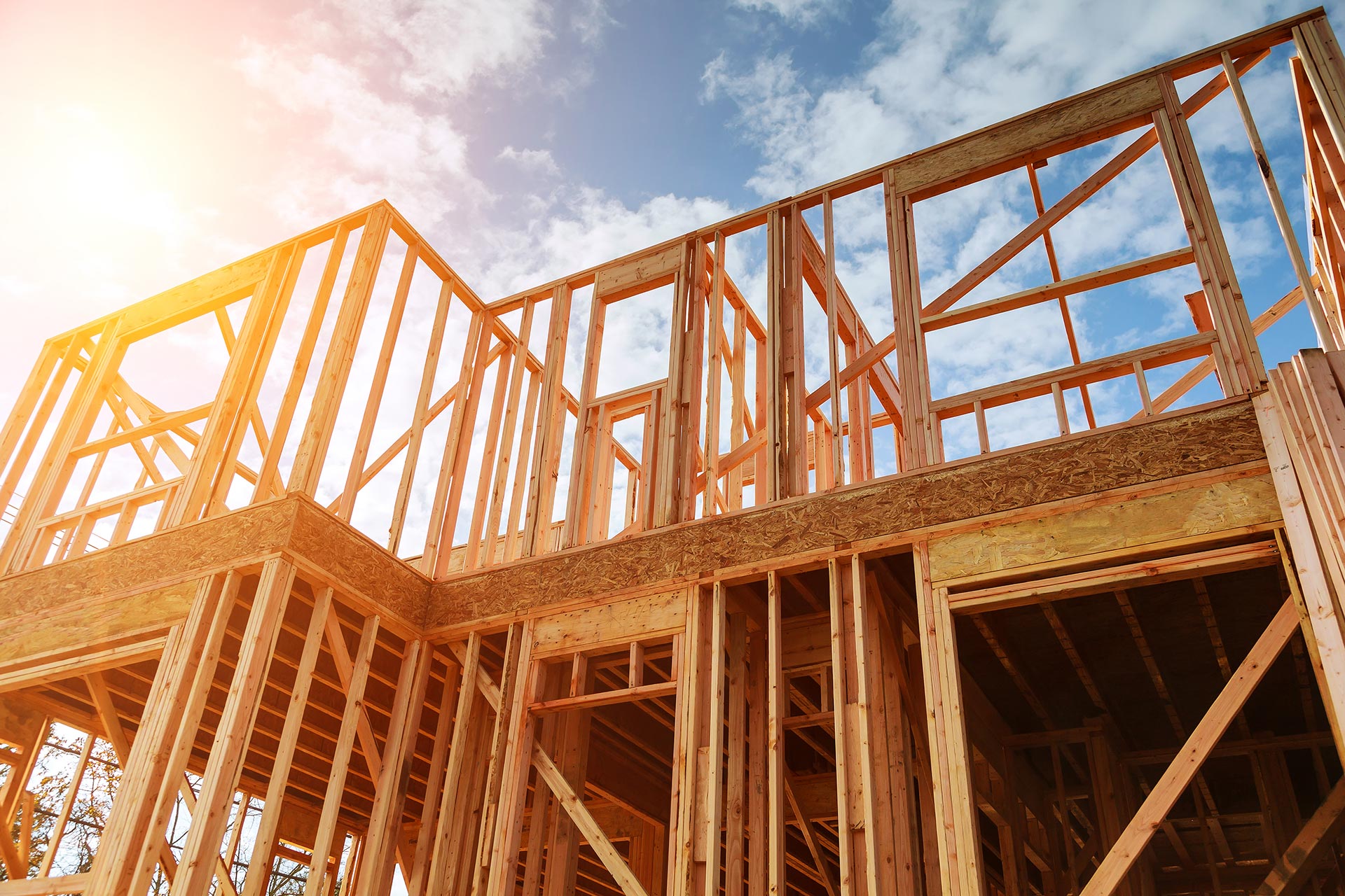 Low-angle view of the structural wood framing of a new two-story home under construction against a bright blue sky with a sun flare. The image showcases professional new construction services, highlighting the initial stages of building a custom home.